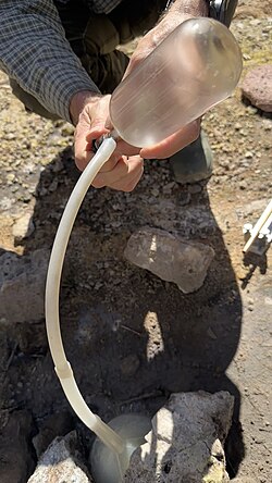 Sampling of noble gases using a Giggenbach bottle, a funnel is placed on top of the hot spring to focus the stream of sample towards the bottle via the Tygon tube. A geochemist is controlling the flow of the sample inlet using a Teflon valve. Note the condensation process inside the evacuated Giggenbach bottle.