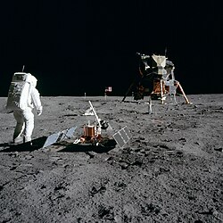 Tranquility Base:[1] (L to R) Astronaut Buzz Aldrin, scientific equipment, Apollo Lunar Module Eagle; US flag and television camera in back (photo by Neil Armstrong)
