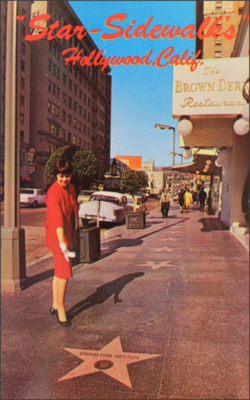 A woman in a red dress points at Charlton Heston's star with other stars, Hollywood Brown Derby, Vine Street, and the Dyas Building viewable behind it. The text "Star-Sidewalks / Hollywood, Calif." printed in a red cursive font at the top of the image
