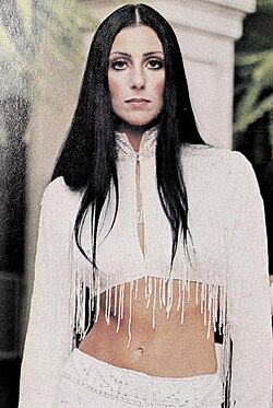 Cher stands in a white, fringed crop top with long sleeves and a white skirt, her long black hair parted in the middle and her midriff exposed.