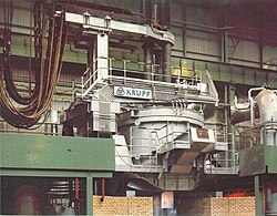 A general overview of a typical AC (Alternating Current) electric arc furnace with three electrodes. All relevant hardware (except the transformer vault to left) are visible, such as water-cooled power cables, painted copper electrode arms, swing roof, refaractory-lined cylindrical melting hearth, tap spout, tilting mechanism, and exhaust ducting.