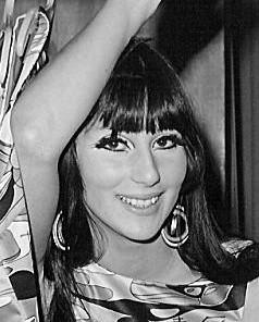 Cher smiles with her arm raised, wearing large hoop earrings and a patterned outfit, surrounded by bold geometric-patterned hanging garments.