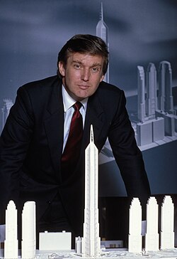 Donald Trump, in a black suit and red tie, standing in from of a model of Manhattan development projects