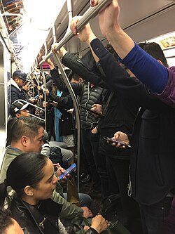 The Q train filled with commuters, many within one inch of each other. Several commuters are seen using smartphones; others are holding on to the train while standing.