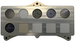 SHERLOC's calibration target aboard the Perseverance Mars rover with Mars Meteorite in the center of the top row and the trackable in the bottom left corner