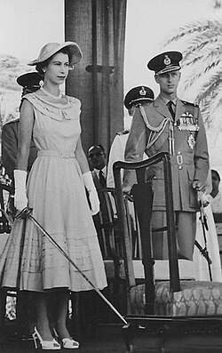 Queen Elizabeth II holding a sword, prepared to knight subjects in Aden as part of a 1954 visit. Prince Philip, Duke of Edinburgh is at right.