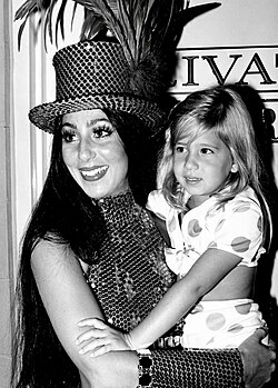 Cher, wearing a sequined top hat with feathers and a matching halter top, smiles while holding young Chaz Bono in a polka-dot outfit.