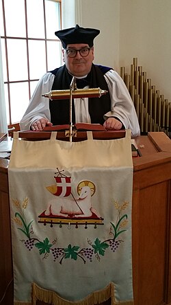 An Anglican minister delivering a homily, dressed in choir habit with Canterbury cap