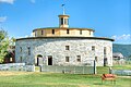 This color photo shows a round barn made of fieldstone. It is topped by a mustard yellow wood frame multi-sided section that is smaller, and then a small turret.