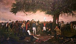 A group of men is gathered under a large tree. One man lays on the ground under the trees, with his barefoot exposed.