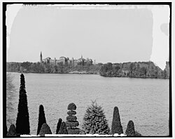 A view of Wellesley College (background) in 1904 from across Lake Waban, with the topiary hedges of the Hunnewell Estate and Gardens in the foreground.