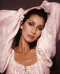 Cher with arms raised above her head, wearing a blush pink peasant blouse with silver leaf embroidery and billowing sleeves, gazing softly at the camera.