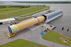 A large orange cylindrical tank pair for the Space Launch System rocket's first stage, being loaded into a long covered barge, on a brown canal
