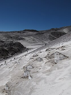Blue-ish ice emerges from underneath grey sand on a mountain slope.