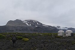 Person walking across rocky ground toward two small shelters, with a mountain in the background under a cloudy sky
