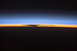 The ozone layer visible from space at Earth's horizon as a blue band of afterglow within the bottom of the large bright blue band that is the stratosphere, with a silhouette of a cumulonimbus in the orange afterglow of the troposphere.