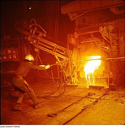 Foaming slag pours out the slag door of an EAF, while a worker checks the temperature using a long probe. Circa 1976
