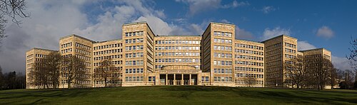 Panoramic view of a massive light-yellow stone building with symmetrical wings, rows of rectangular windows, and a central entrance under a cloudy blue sky