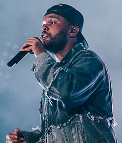 A slight worm's eye-shot of the Weeknd, with a black cap worn backwards and a blue jacket, performing while holding a microphone.