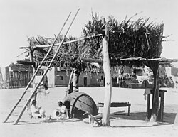 A Zuni drying platform for maize and other foods, with two women crafting pottery beneath it. From the Panama-California Exposition, San Diego, California. January 1915.