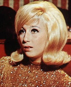 Cher with a voluminous blonde bouffant hairstyle and winged eyeliner, wearing a gold sequined top while seated in a red booth.