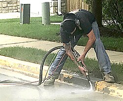 Bead blasting paint from a concrete curb by a worker wearing hearing protection. Mixing particles with water substantially reduces dust.