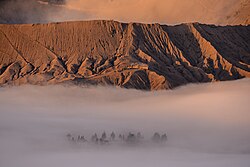 The steps leading to the rim of Mount Bromo's crater are clearly visible; below them lies the Pura Luhur Poten Bromo temple, shrouded in mist.