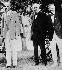 Black and white photo taken in bright light shows three men in suits standing in a grassy field surrounded by a few large trees. Edison appears more elderly.
