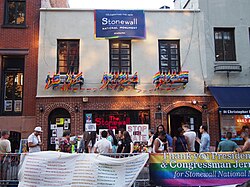 A two-story building with brick on the first floor, with two arched doorways, and gray stucco on the second floor, off of which hang numerous rainbow flags.