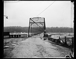 The Potomac River surges over the deck of Chain Bridge during the historic 1936 flood. The bridge was so severely damaged by the raging water, and the debris it carried, that its superstructure had to be re-built; the new bridge was opened to traffic in 1939. (This photograph was taken from a vantage point on Glebe Road in Arlington County, Virginia. The houses on the bluffs in the background are located on the Potomac Palisades of Washington, DC.)