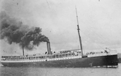 A grainy black and white photo of a steamship on the water. The bow faces to right and the ship occupies the whole frame. Several groups of people are visible fore and aft, on the first and second deck. The ship has two masts and a smokestack which is bellowing.